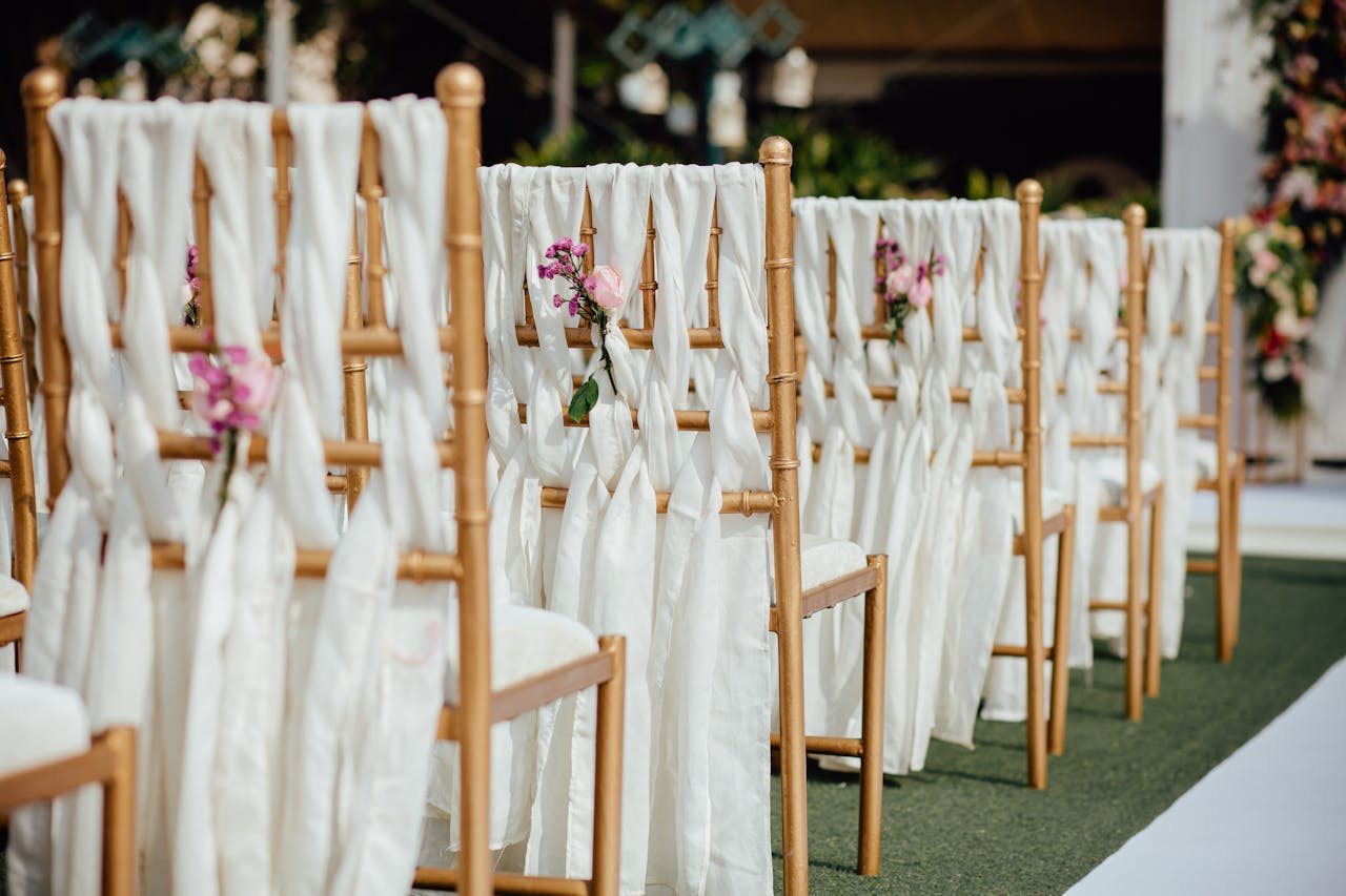 Row of elegant wooden chairs decorated with white fabric and pink flowers for an outdoor wedding ceremony.